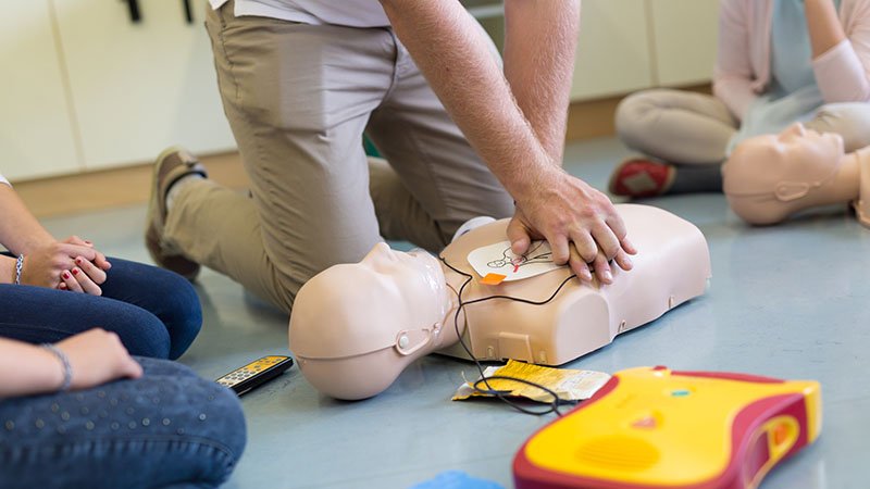 En person demonstrerer hjerte‑ og lungeredning på en treningsdukke med en hjertestarter, mens flere sitter og følger med.