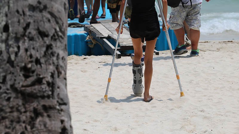 En dame går i sanden med krykker og en gips på beinet