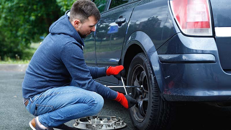 Mann bytter dekk på en bil og strammer hjulbolter med en hjulnøkkel, mens hjulkapselen ligger på bakken ved siden av.