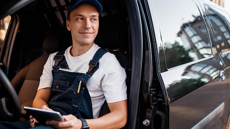 Person i arbeidsklær sitter i førersetet på en bil, med døren åpen og holder et nettbrett.