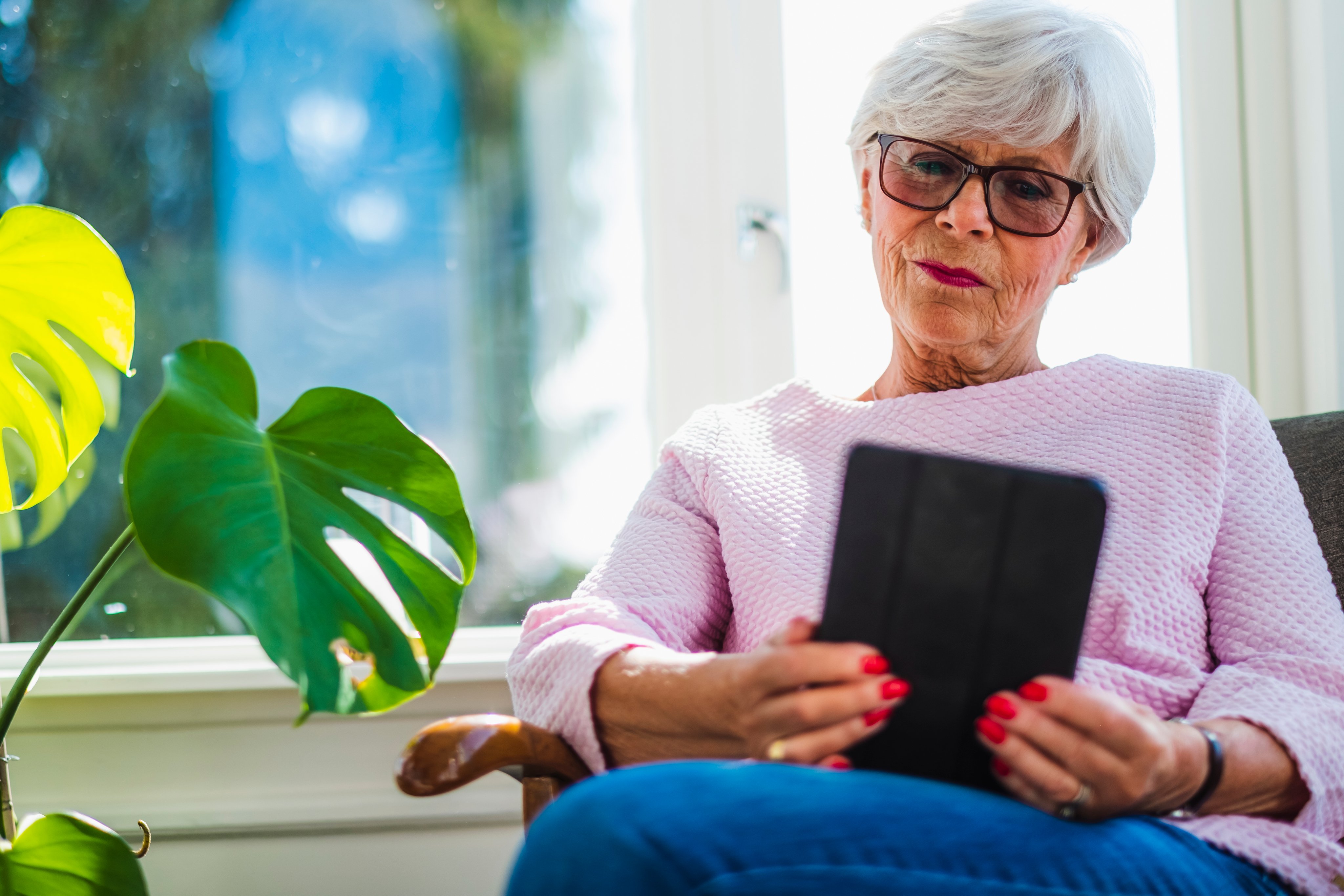 En eldre kvinne sitter i en stol og leser på et nettbrettet.