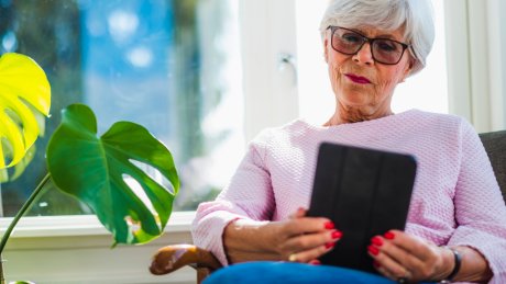 En eldre kvinne sitter i en stol og leser på et nettbrettet.