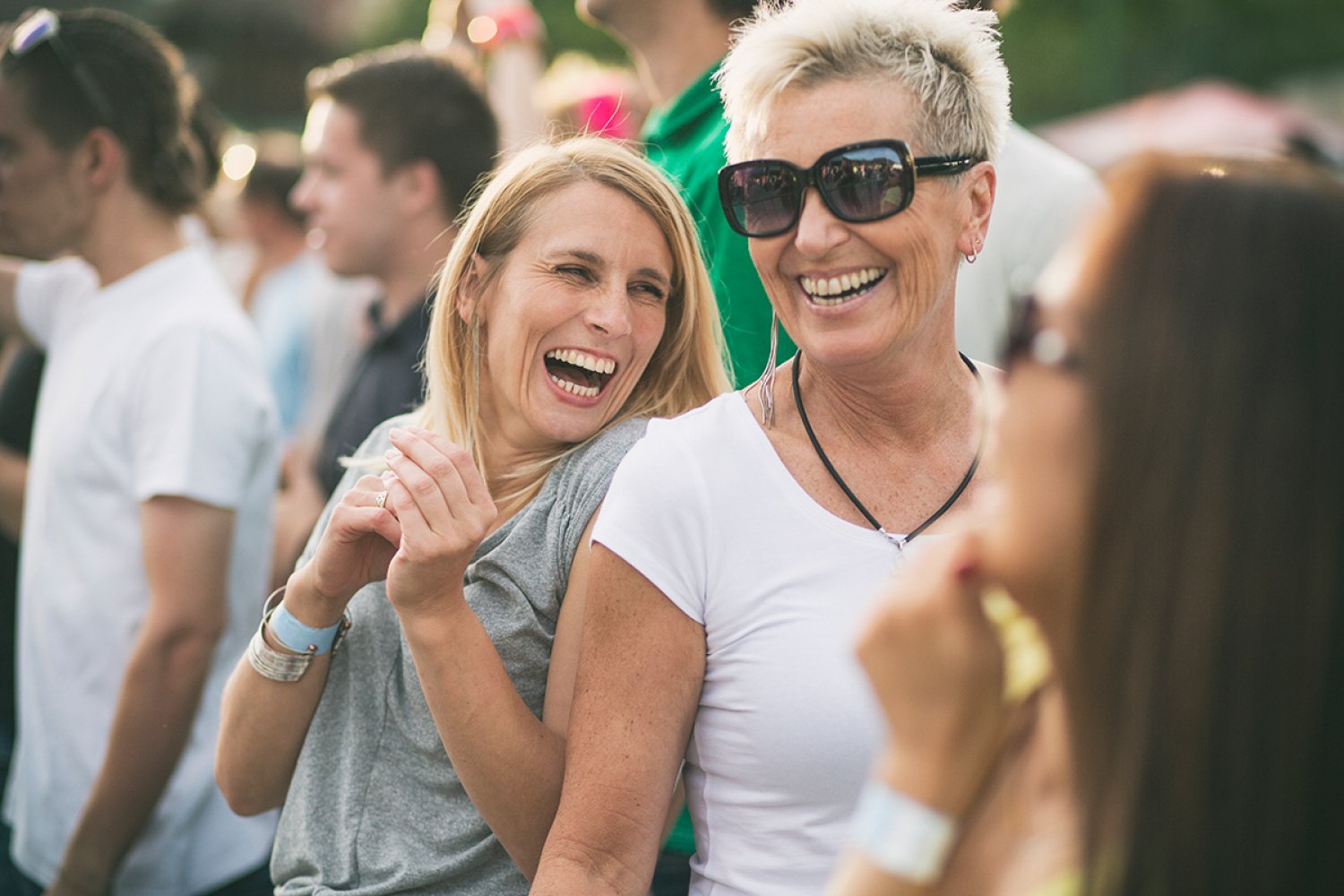 To eldre damer smiler og ler på en utendørskonsert i sola.