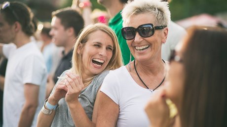 To eldre damer smiler og ler på en utendørskonsert i sola.