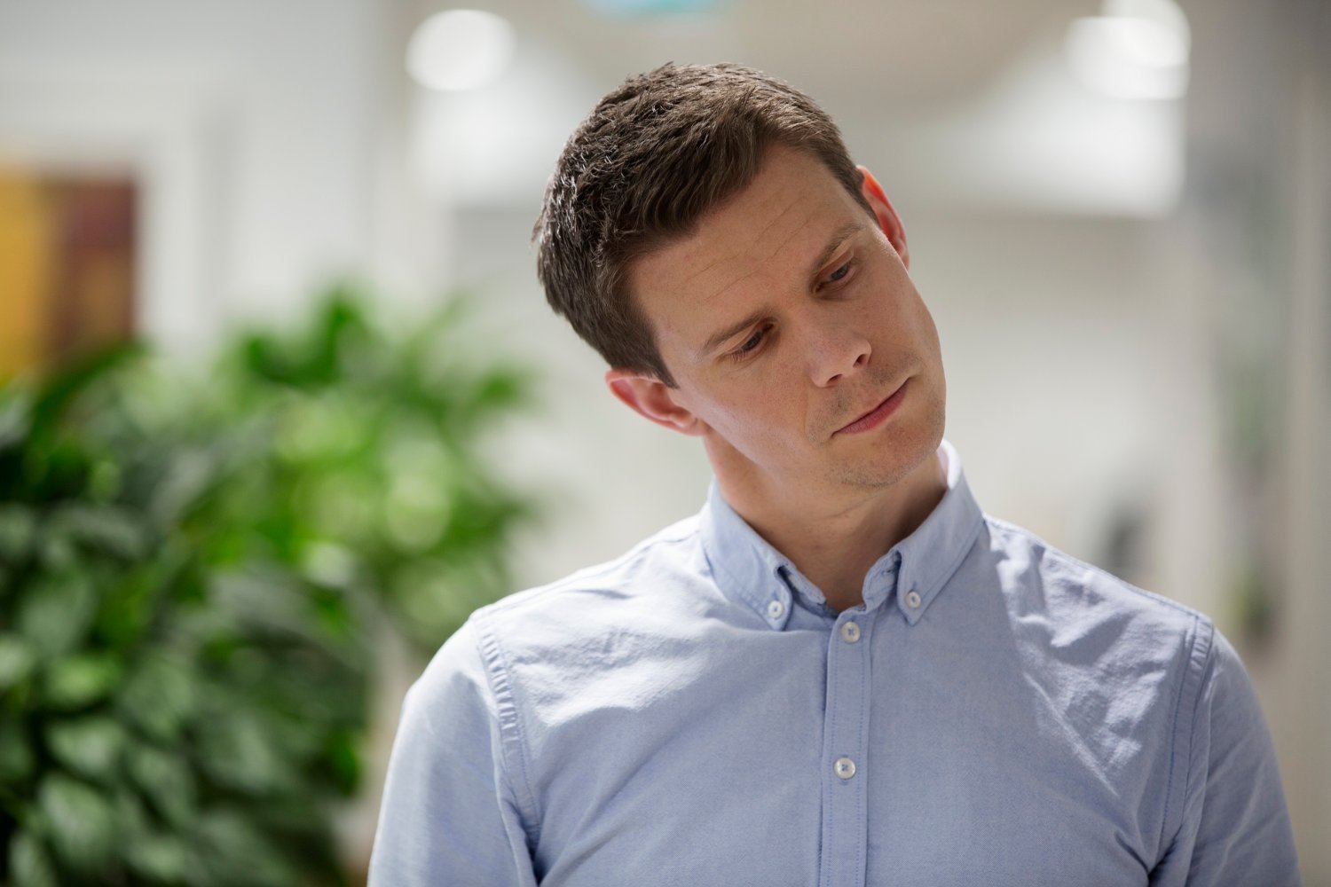 Person med lyseblå skjorte står innendørs i et kontormiljø med grønne planter og lys bakgrunn.