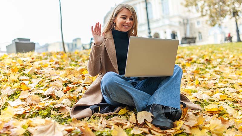 Kvinne sitter i en park med en laptop i fanget, vinker mot laptoppen.