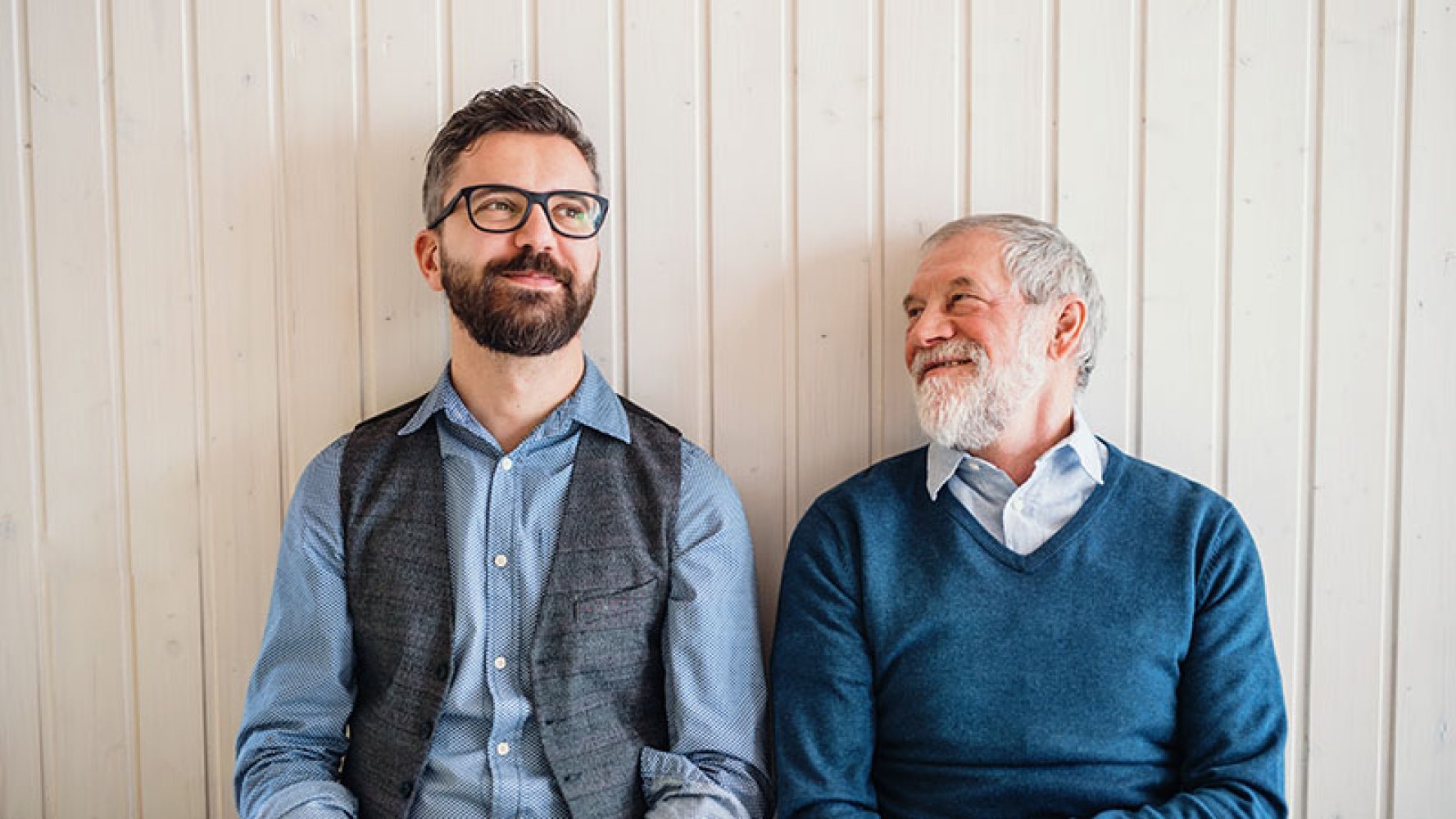 To menn sitter side ved side og smiler varmt til hverandre foran en lys trevegg.