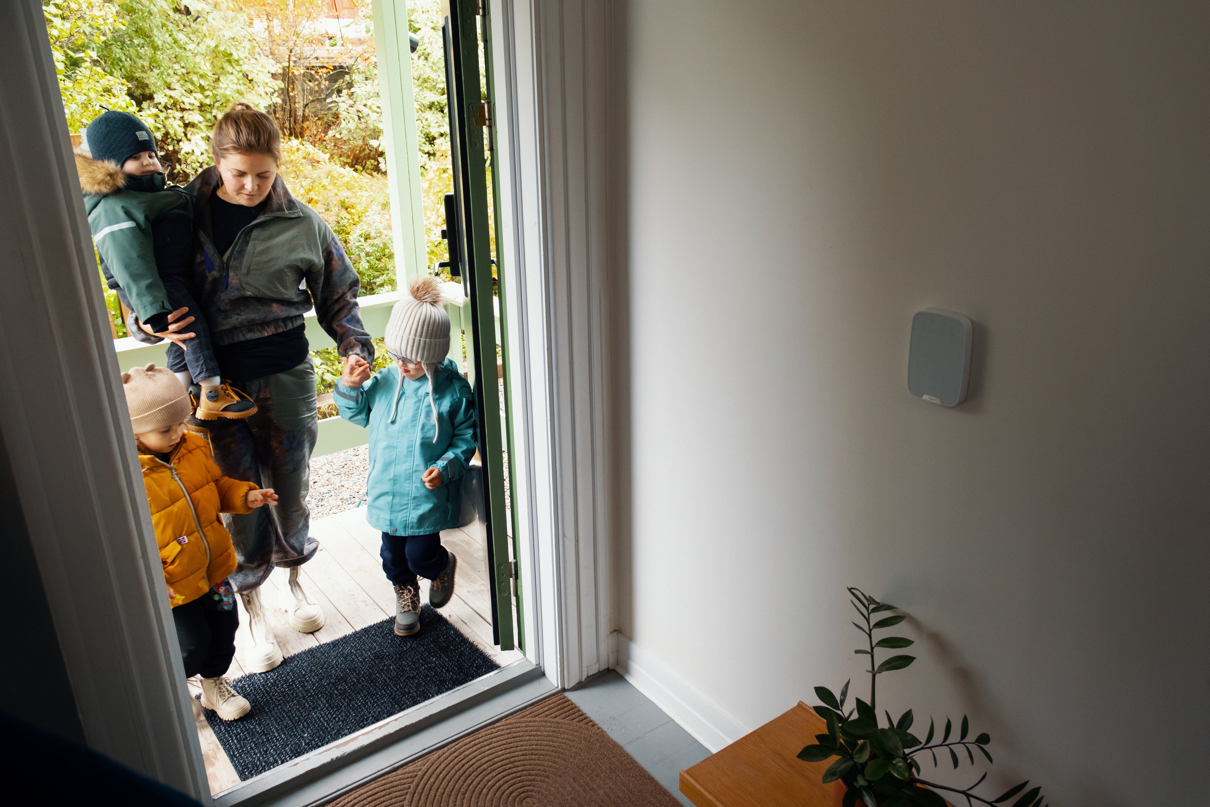 Mor med tre små barn på vei inn inngangsdør i en bolig, kledd i turtøy. På veggen i gangen på huset er montert et Smart Hus-kontrollpanel. 