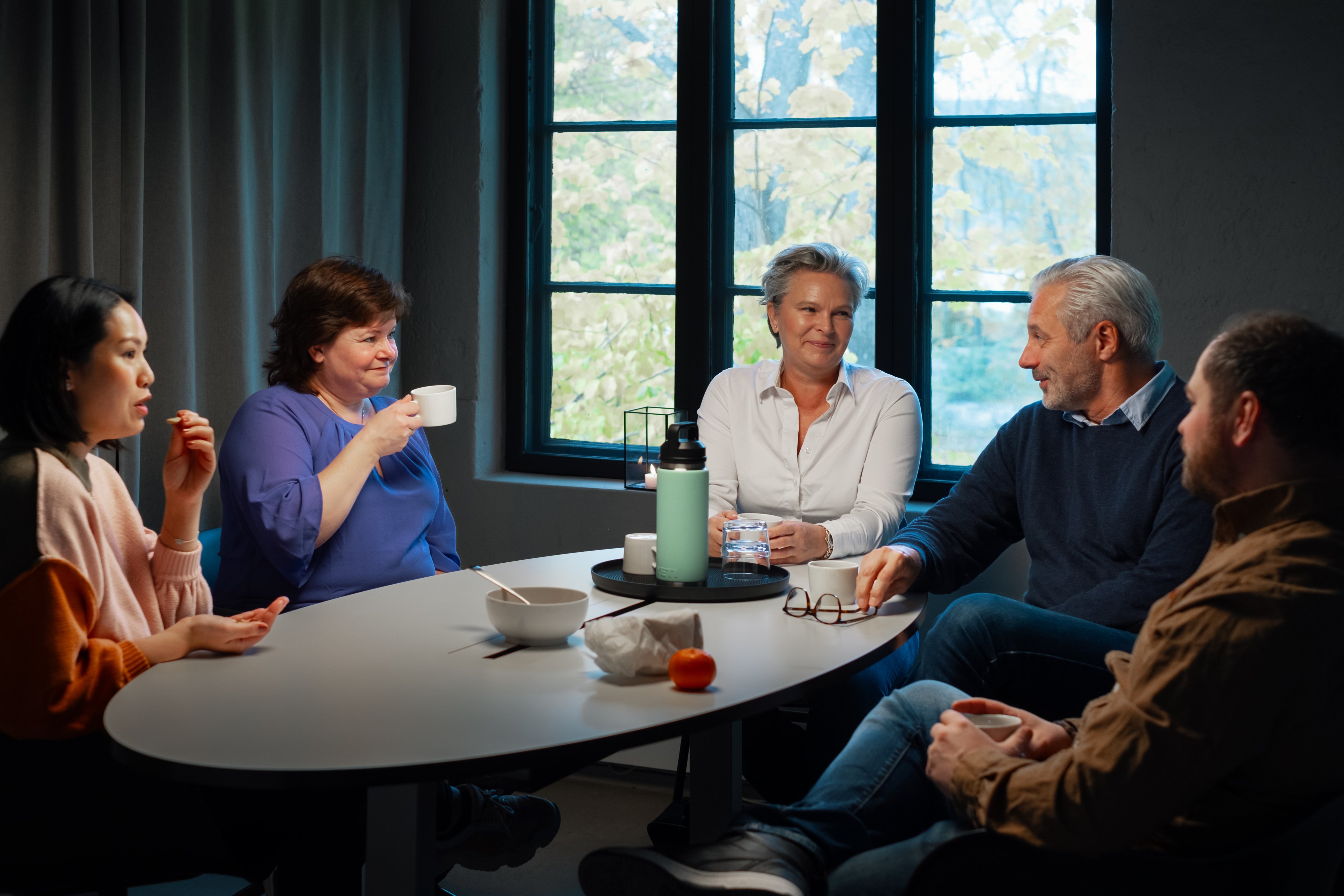 Fem kollegaer sitter rundt et bord og prater og drikker kaffe,