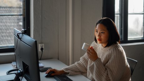 Kvinne ved skrivebord med kaffekopp foran dataskjerm.