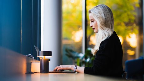 Ung kvinne sett fra siden som sitter og jobber på PC ved et bord