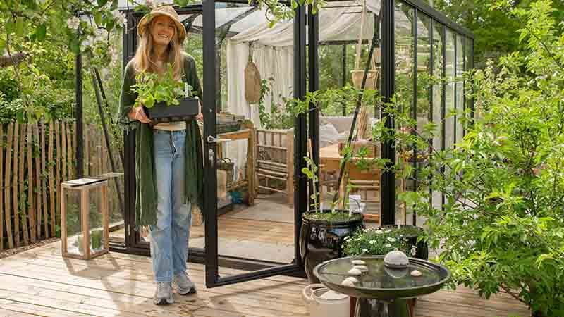 En kvinne står foran et drivhus med en plante i armene.