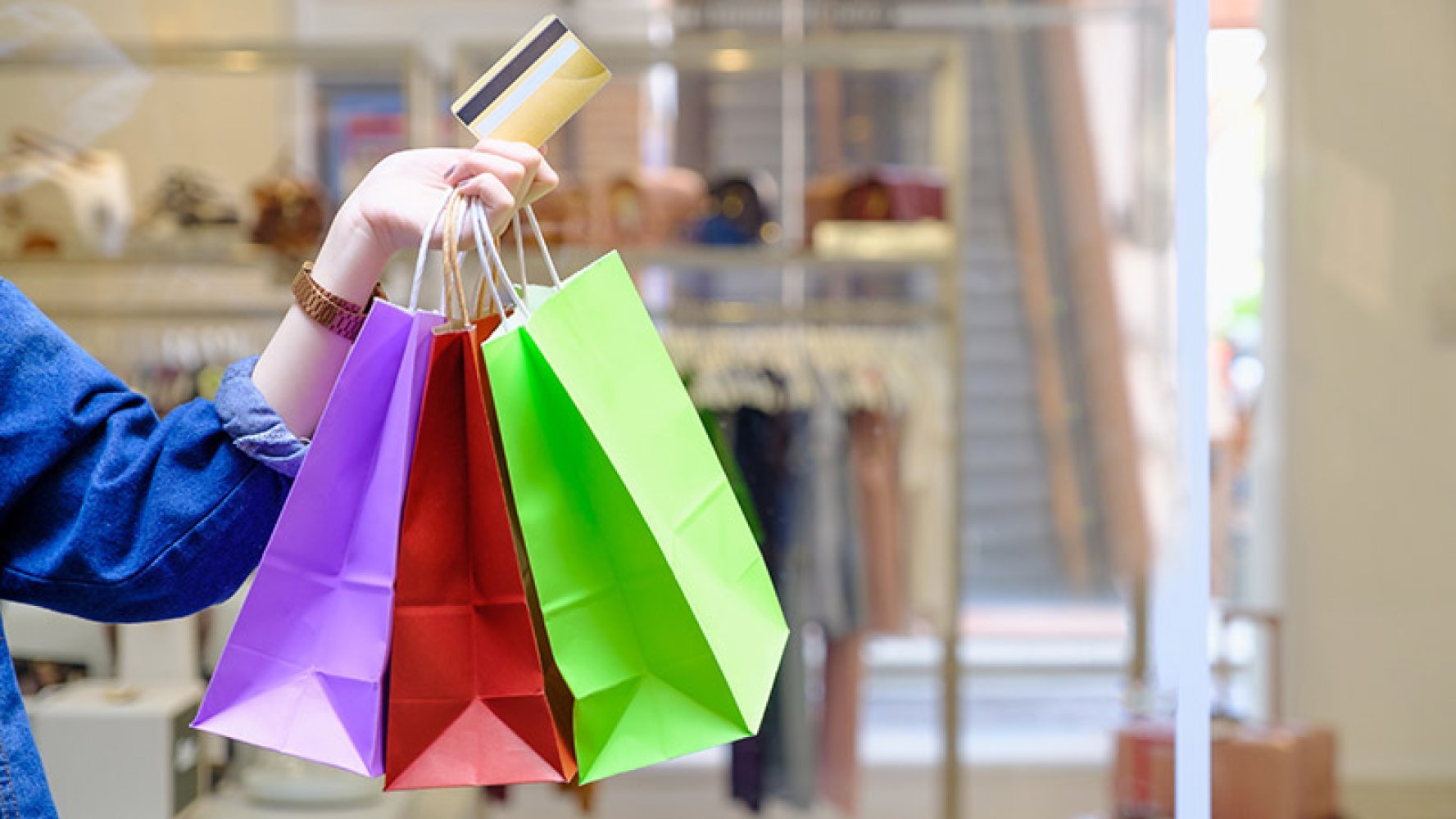 Hånd som holder flere fargerike handleposer og et kredittkort i et kjøpesenter, med klesstativer og butikkinteriør i bakgrunnen.