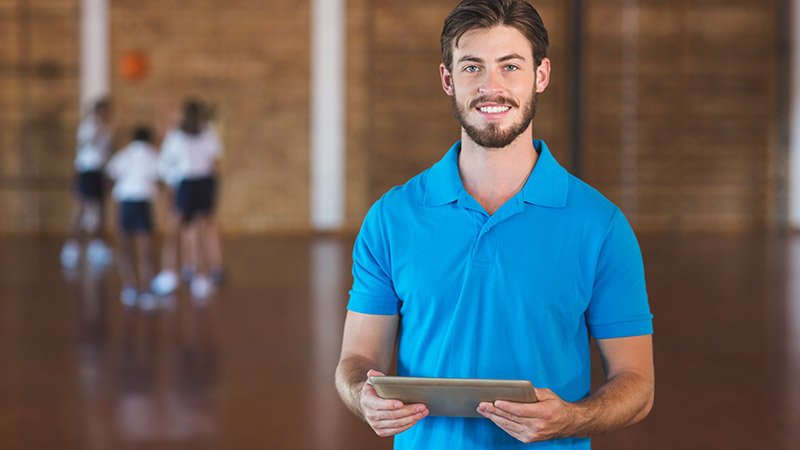 Mann i blå poloskjorte holder et nettbrett i en gymsal, mens elever spiller i bakgrunnen.