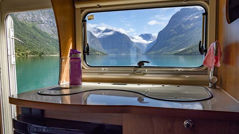 Innsiden av en campingvogn, og gjennom et vindu og en åpen dør ser man et blått vann og dramatisk fjellandskap.