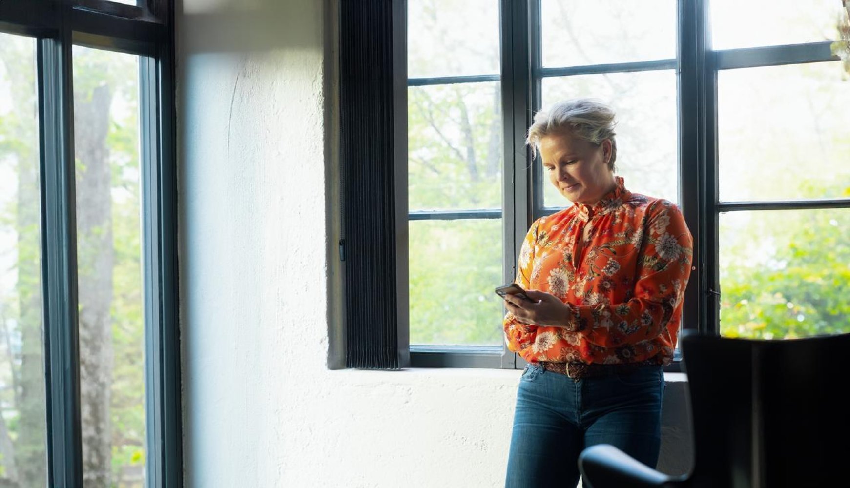 En dame står foran et vindu innendørs og ser på mobiltelefonen.