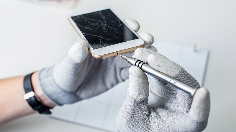 En person med hansker bruker et presisjonsverktøy for å reparere en mobiltelefon med knust skjerm.