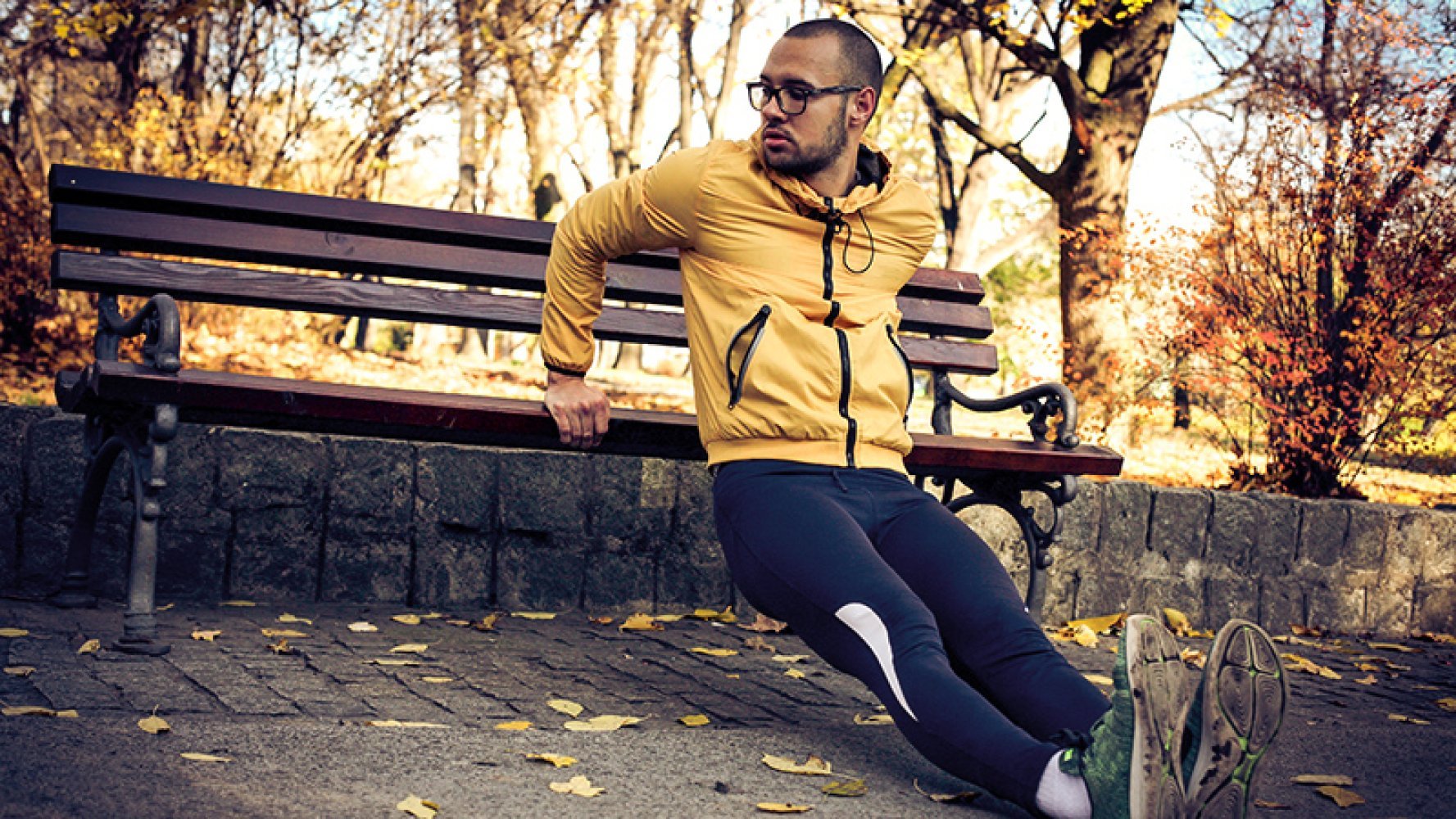 Ung mann i treningsklær gjør "dips" på en benk ute i solfylt høstværet
