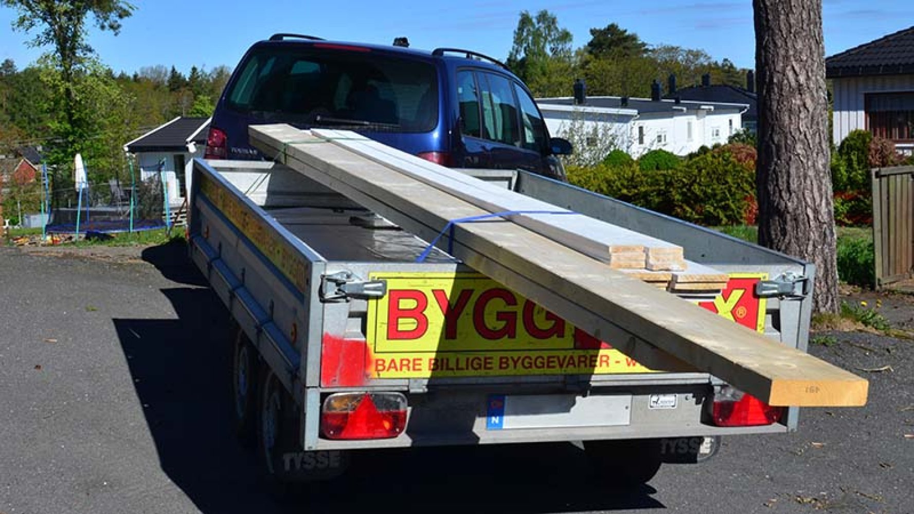 Biltilhenger festet til bil, med flere planker liggende oppå karmen til hengeren, festet med tau.