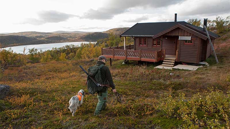 Person med jaktutstyr og hund går mot en hytte i fjellet, med innsjø og høstfarger i bakgrunnen.