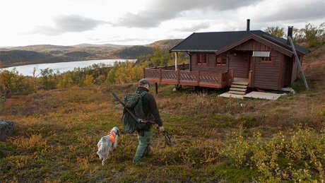Person med jaktutstyr og hund går mot en hytte i fjellet, med innsjø og høstfarger i bakgrunnen.
