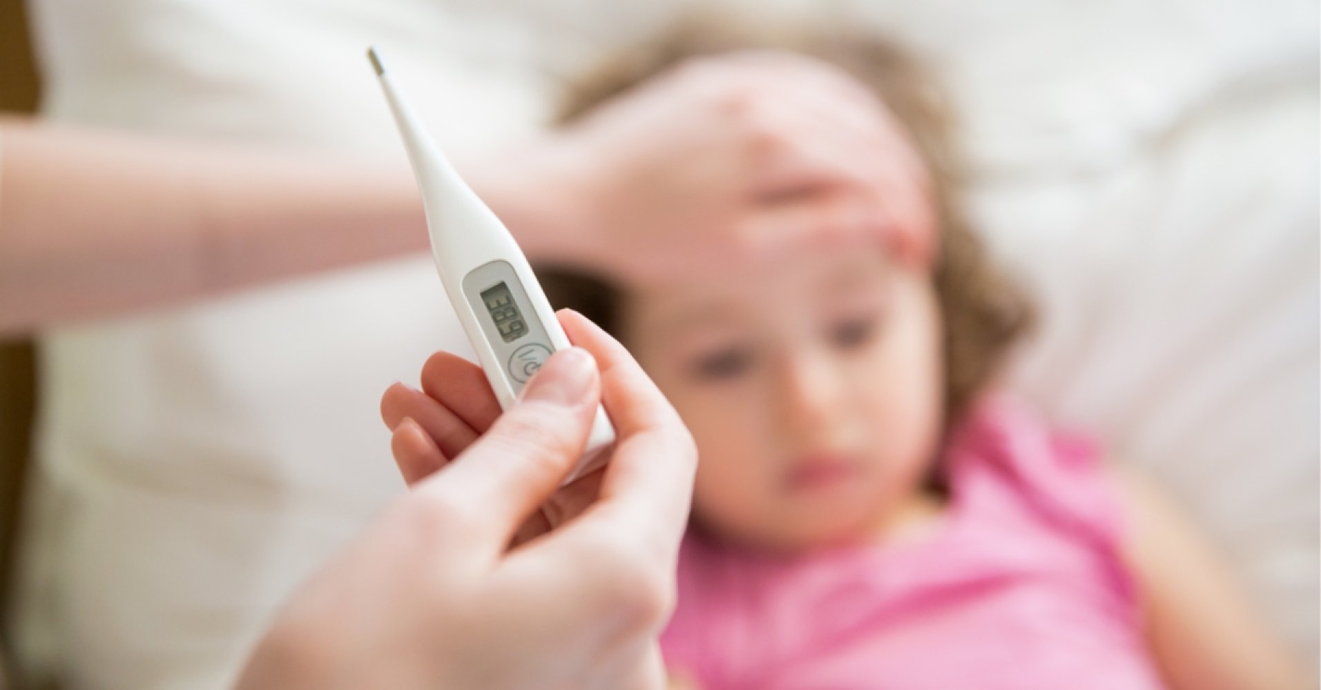 Termometer som viser 38,9 grader i fokus, med et sykt barn med en hånd på pannen i bakgrunnen