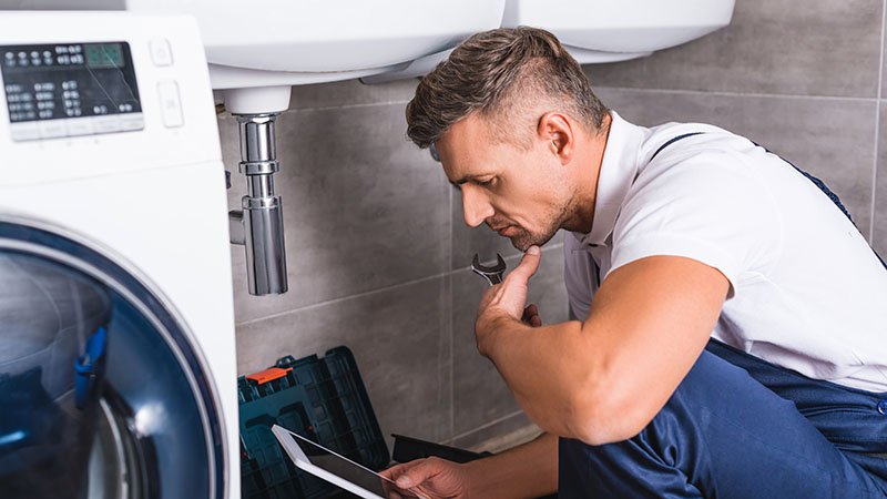 En rørlegger sitter på gulvet foran et åpent verktøyskrin og undersøker rørene under en vask, med et nettbrett i hånden. Til venstre står en vaskemaskin.