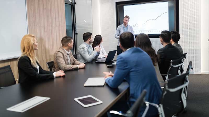 Åtte medarbeidere lytter til en som viser grafer i et møterom.