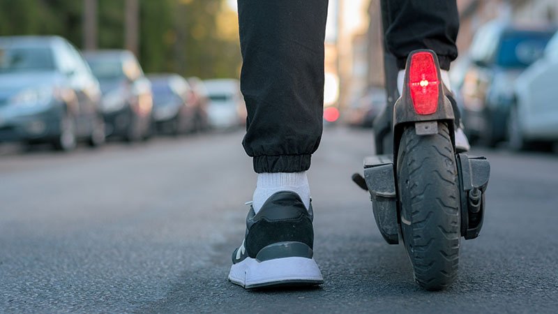 Nærbilde av en person som står på en elsparkesykkel, ser bare personen fra leggen og ned og bakhjulet på elsparkesykkelen.
