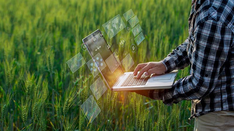 Person i rutete skjorte jobber på laptop i et grønt kornfelt, med transparente data- og kartikoner over skjermen.