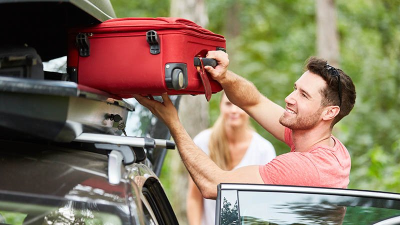 Mann legger en koffert i takboksen på biltaket, omgitt av grønn skog i bakgrunnen.