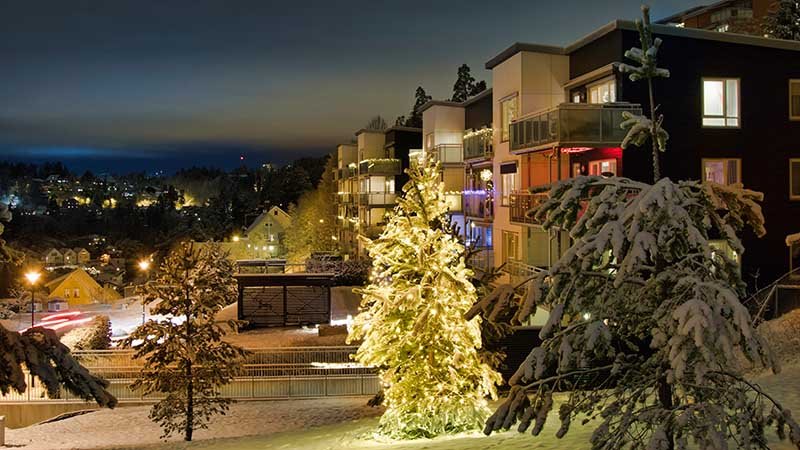 Snødekt boligområde om kvelden med opplyst tre i forgrunnen og leilighetsbygg med balkonger i bakgrunnen.