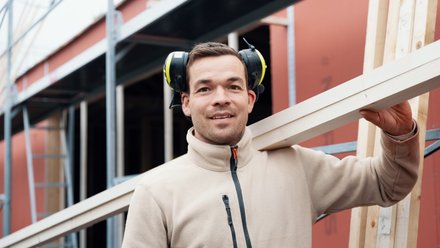 Snekker bærer planker på en arbeidsplass og smiler til kamera