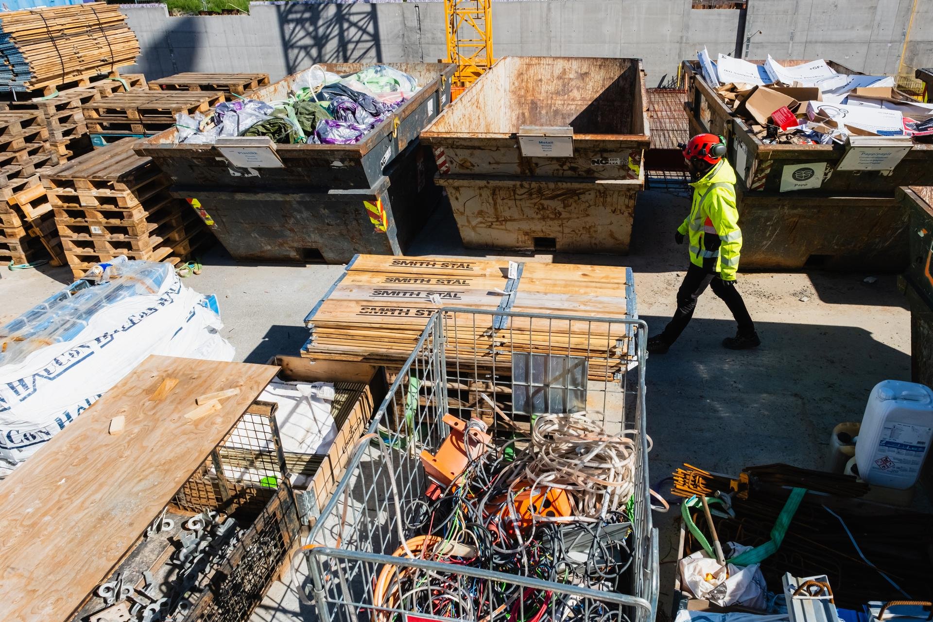 Byggeplass med sorteringsområde: containere fylt med plast og papp, stablede paller og en person i verneutstyr som går forbi.