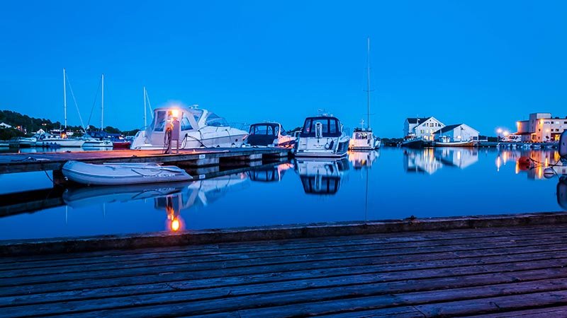 Flere båter ligger fortøyd ved en brygge i stille vann under blå kveldshimmel, med hus og bygninger i bakgrunnen