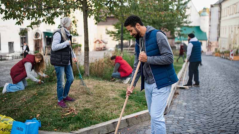 Personer i fritidsklær samarbeider om opprydding i et parkområde. Én raker ved fortauskanten, mens andre plukker avfall i gresset.