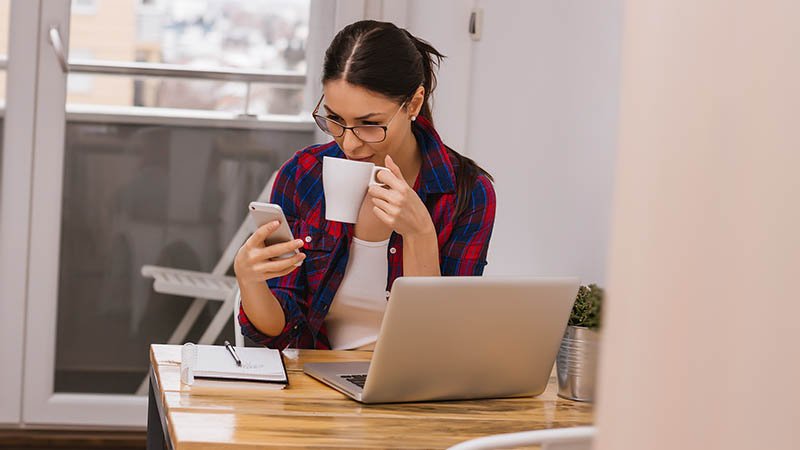 Kvinne sitter ved et lite bord med laptop og skrivebok, drikke fra en kaffekopp og ser på mobiltelefonen sin
