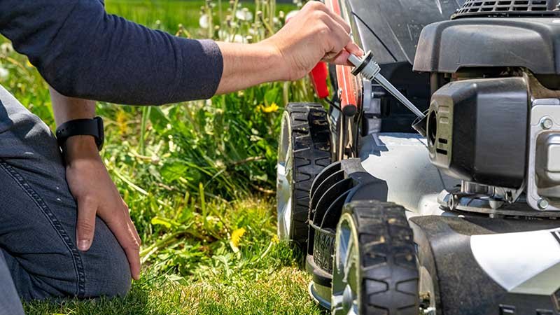 Mann sitter på kne og stikker et redskap ned i oljetanken på gressklipper