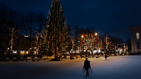 Stor julegran og trær dekorert med lys på en snødekt plass i byen om kvelden, med folk som går gjennom området