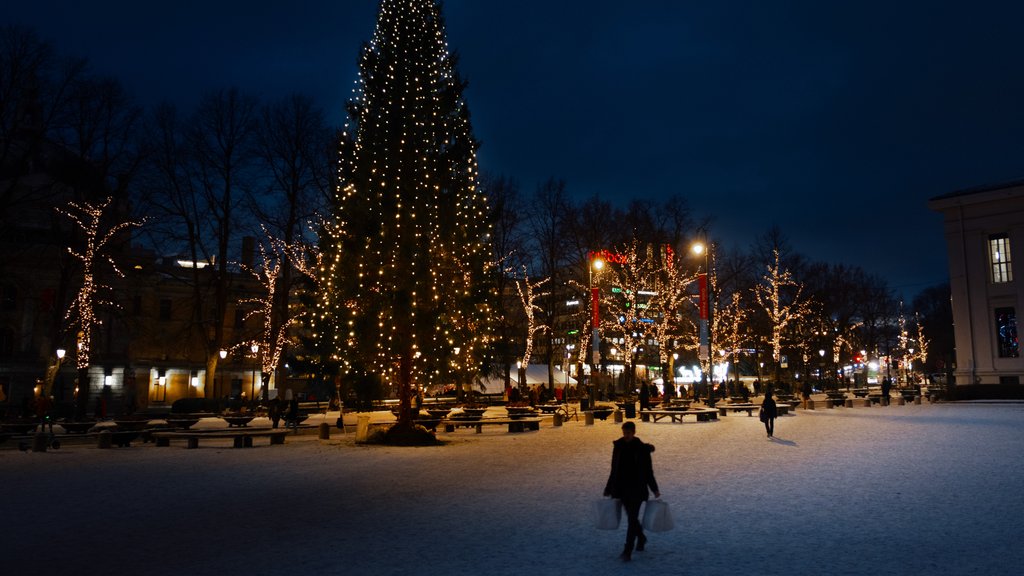 Stor julegran og trær dekorert med lys på en snødekt plass i byen om kvelden, med folk som går gjennom området