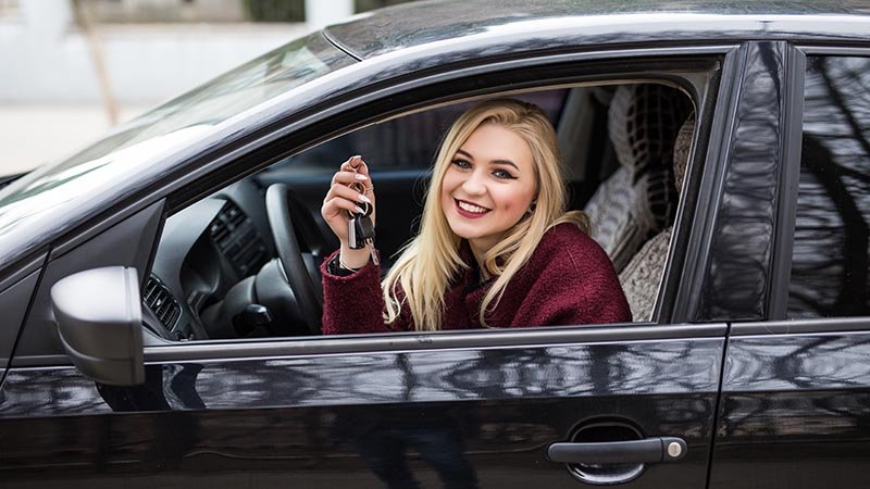 Ung kvinne sitter i en bil og holder  bilnøkler i hånda mens hun smiler.