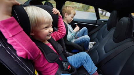 To barn sitter bak i en bil, ett i barnesete og ett i en bilstol.