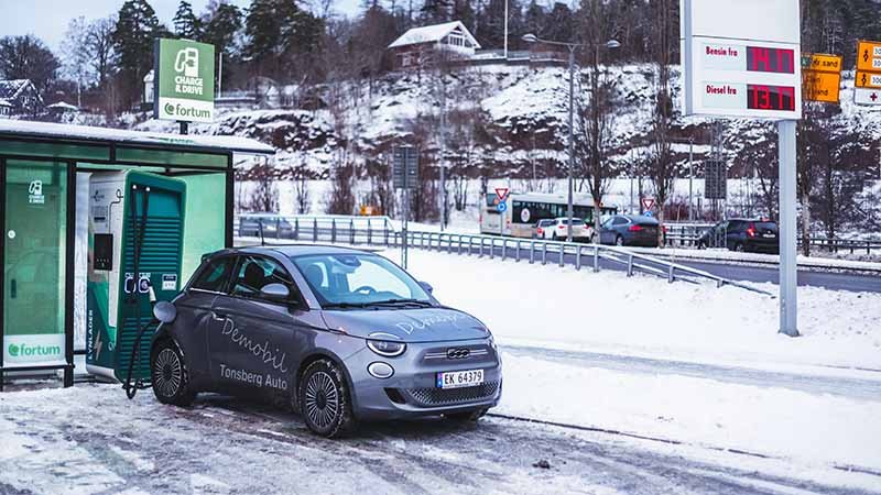 Elbil lader ved en ladestasjon på en snødekt parkeringsplass, med veiskilt og biler i bakgrunnen.