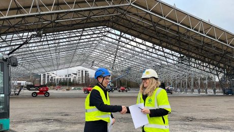 To personer i gule vernevester og hjelmer står på en stor byggeplass med en stålkonstruksjon som dekker området. De håndhilser og holder dokumenter, mens flere maskiner og bygninger er synlige i bakgrunnen.
