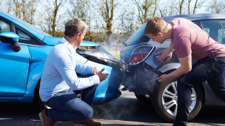 To biler har kollidert, en blå bil har kjørt inn i bakenden på en grå bil, med synlige skader og røyk mellom bilene.