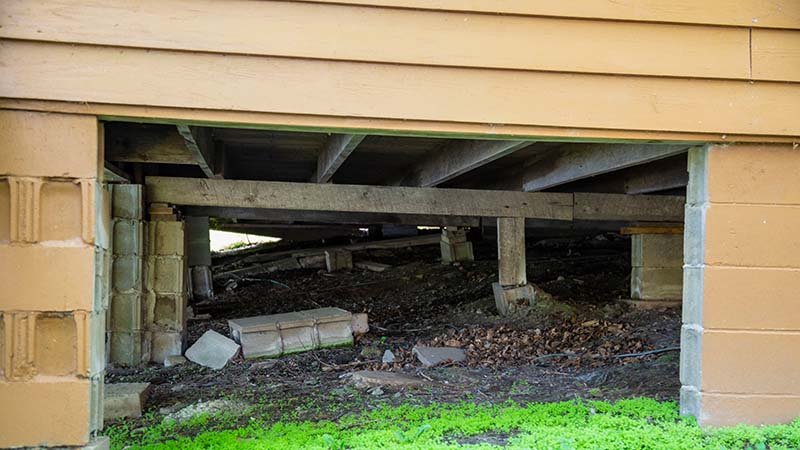 Under et hus med synlige trebjelker og murblokker, der noen blokker er forskjøvet og ligger på bakken.