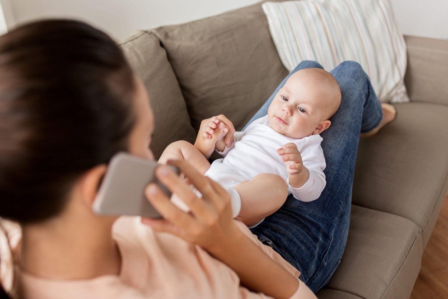 En kvinne sitter i sofaen og holder en baby på fanget mens det snakkes i mobiltelefon.