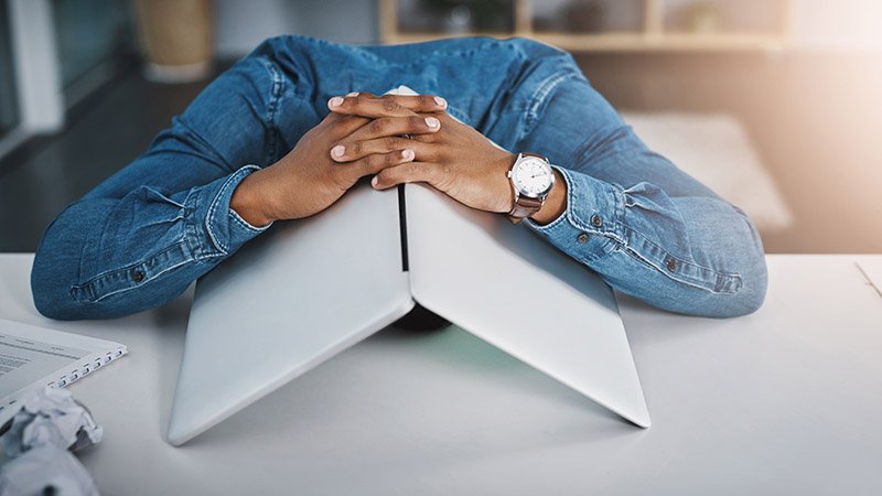 Sliten mann med sammenfoldet laptop over hodet, med hendene foldet oppå, sittende ved et skrivebord.