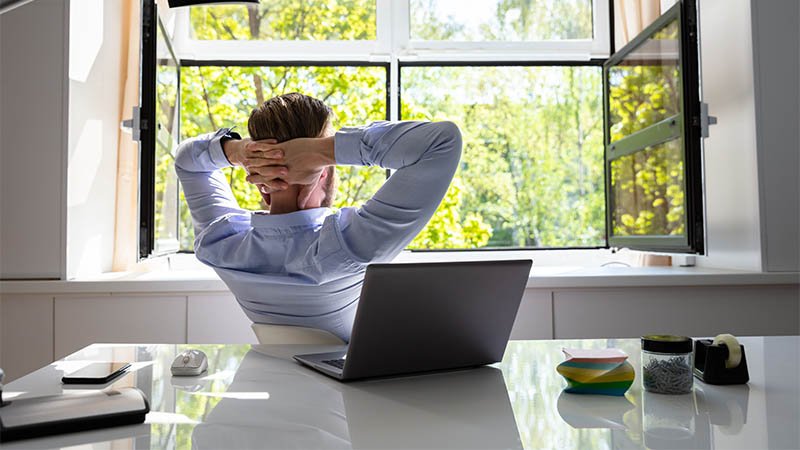 En mann sitter ved et skrivebord med en bærbar PC og ser ut av et åpent vindu mot grønne trær, med hendene foldet bak hodet.