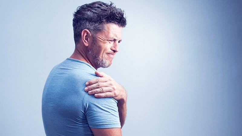 En middelaldrende mann holder hånden på skulderen sin og har et ansiktsuttrykk som viser ubehag.